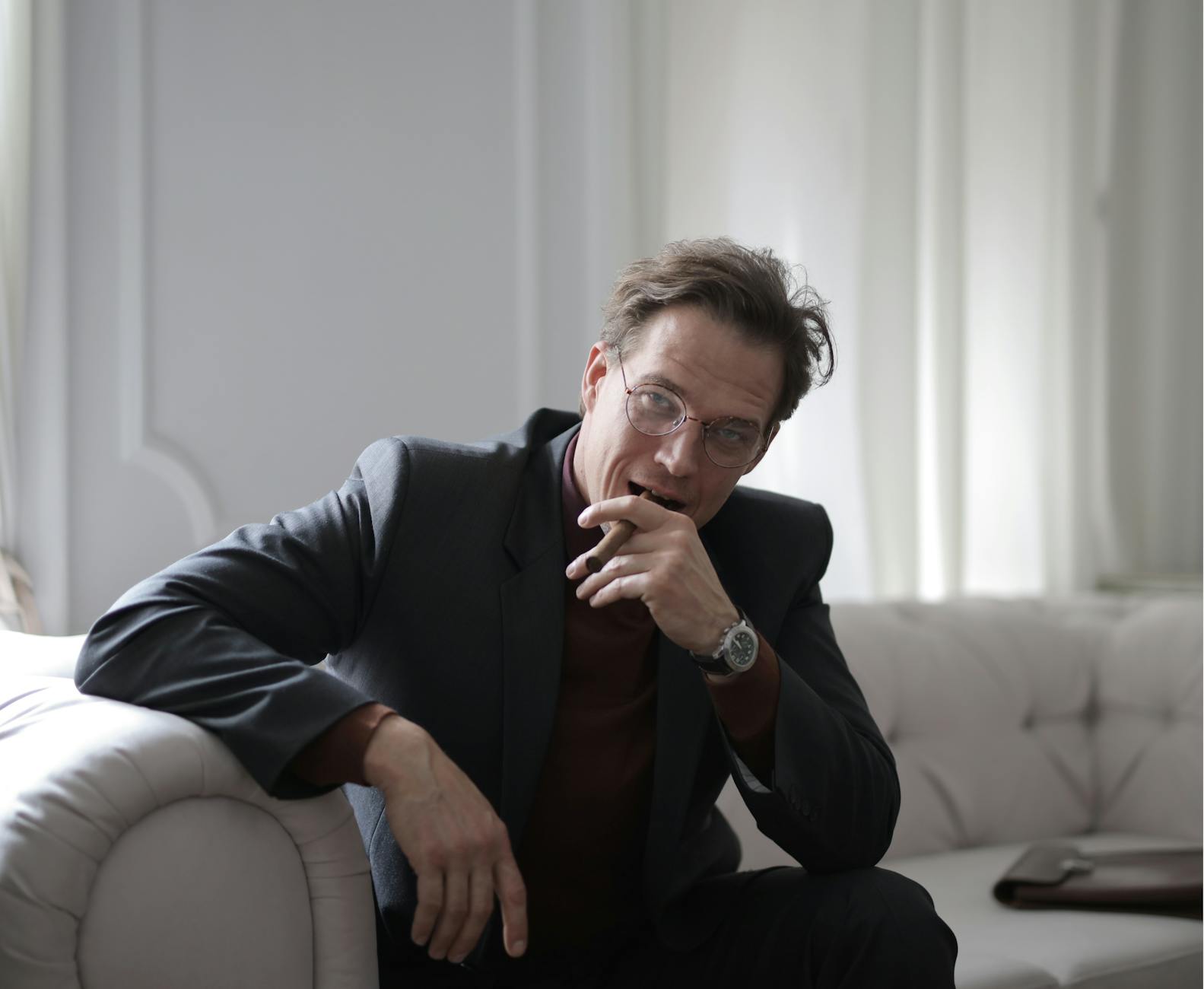 A confident businessman in a black suit sitting on a sofa indoors with a thoughtful expression.
