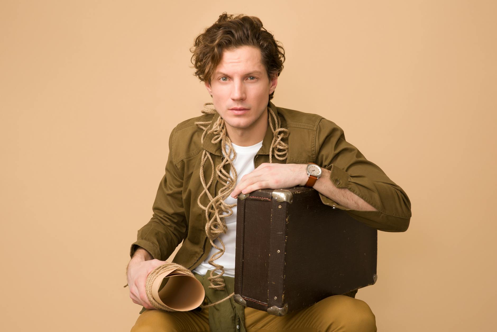 Young man sitting with a vintage suitcase and rope, exuding a stylish and thoughtful pose.