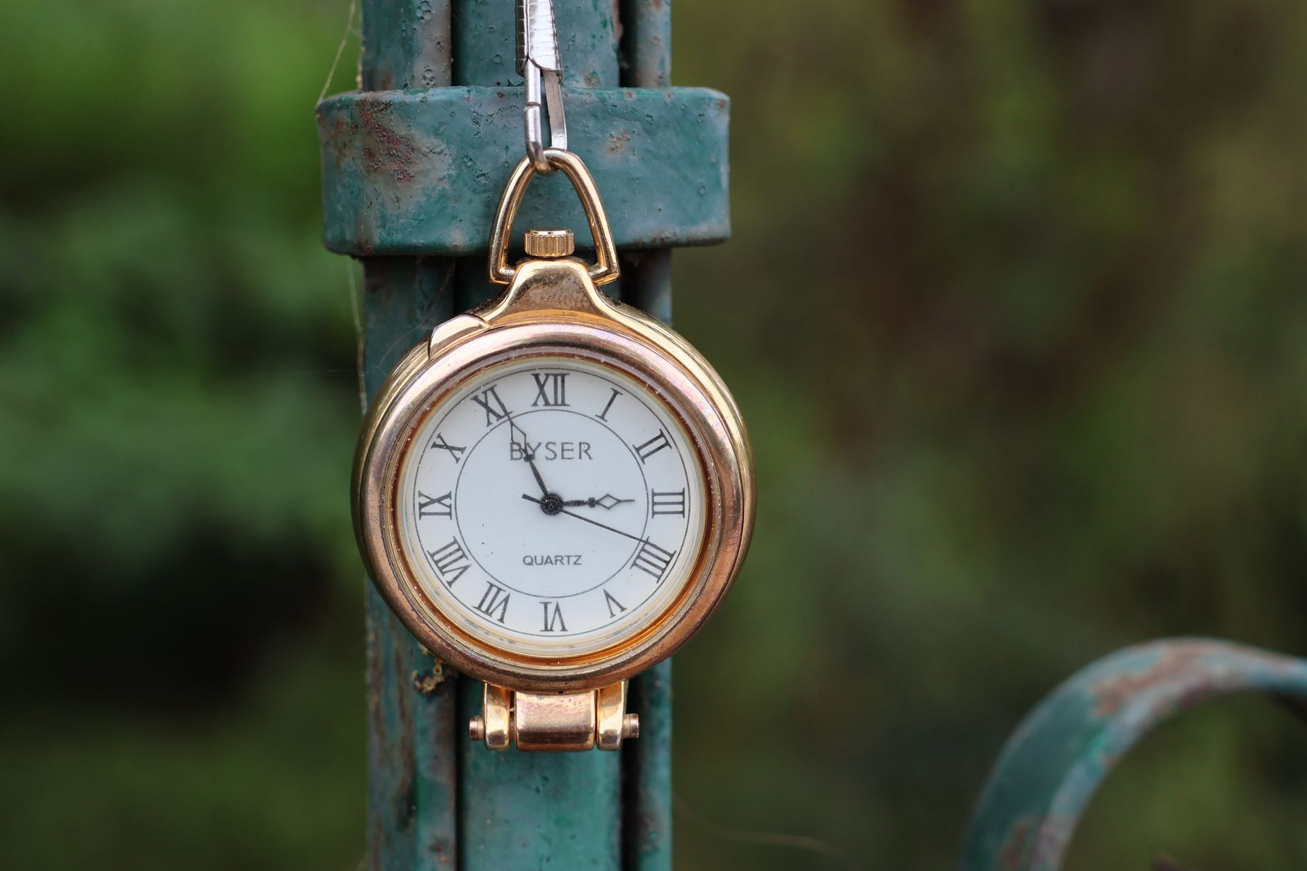 A vintage pocket watch with Roman numerals hanging on a rustic green metal fence outdoors.
