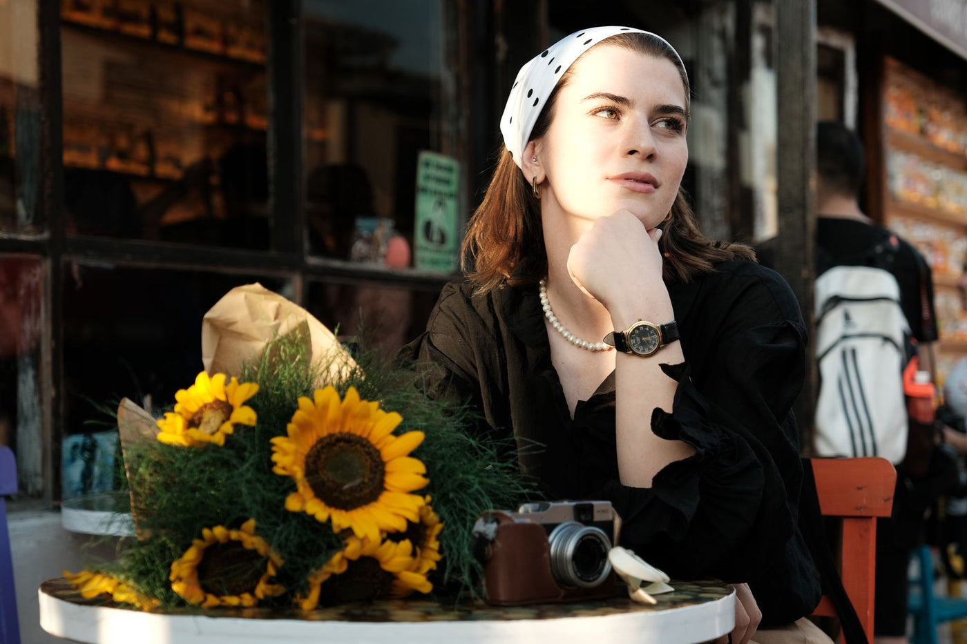 A woman sits by sunflowers at an outdoor cafe, enjoying a sunny afternoon.