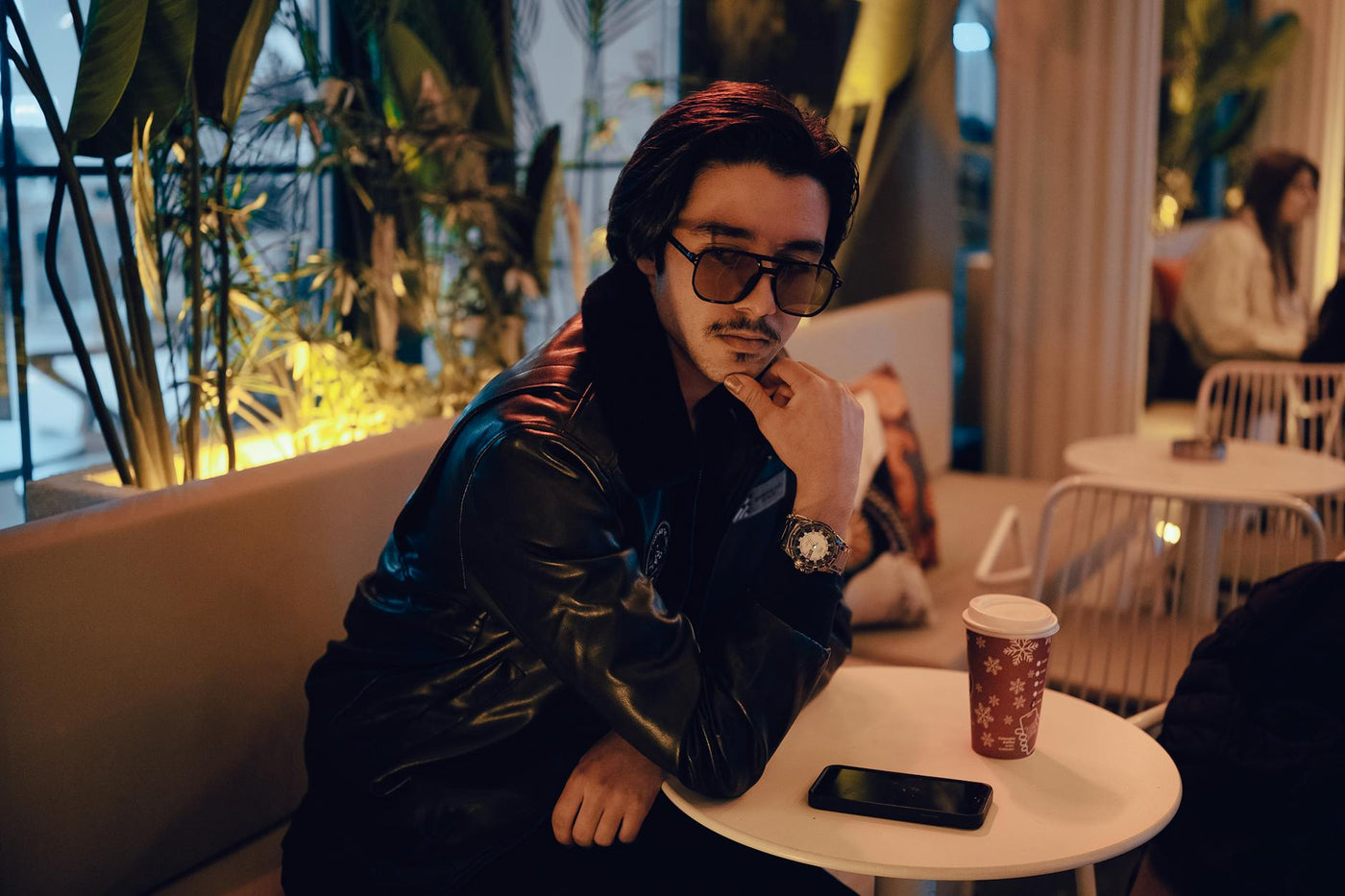 A man in a leather jacket relaxes in a cozy cafe with ambient lighting and a festive coffee cup.