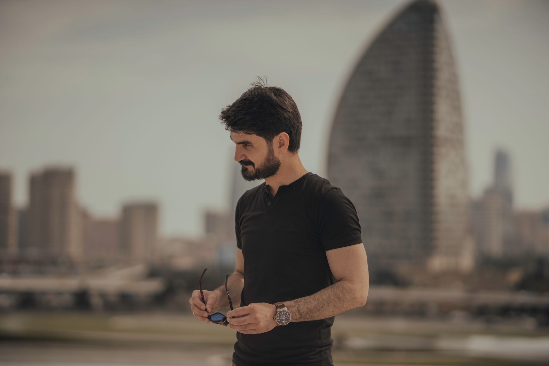 Stylish man in modern cityscape, holding sunglasses with a thoughtful pose.