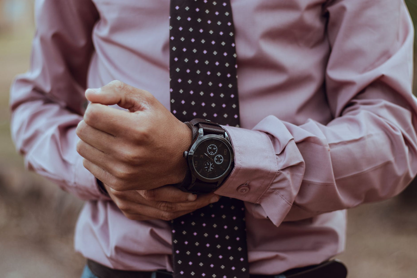 Close-up of an adult man in a pink shirt adjusting his wristwatch and polka dot necktie outdoors.