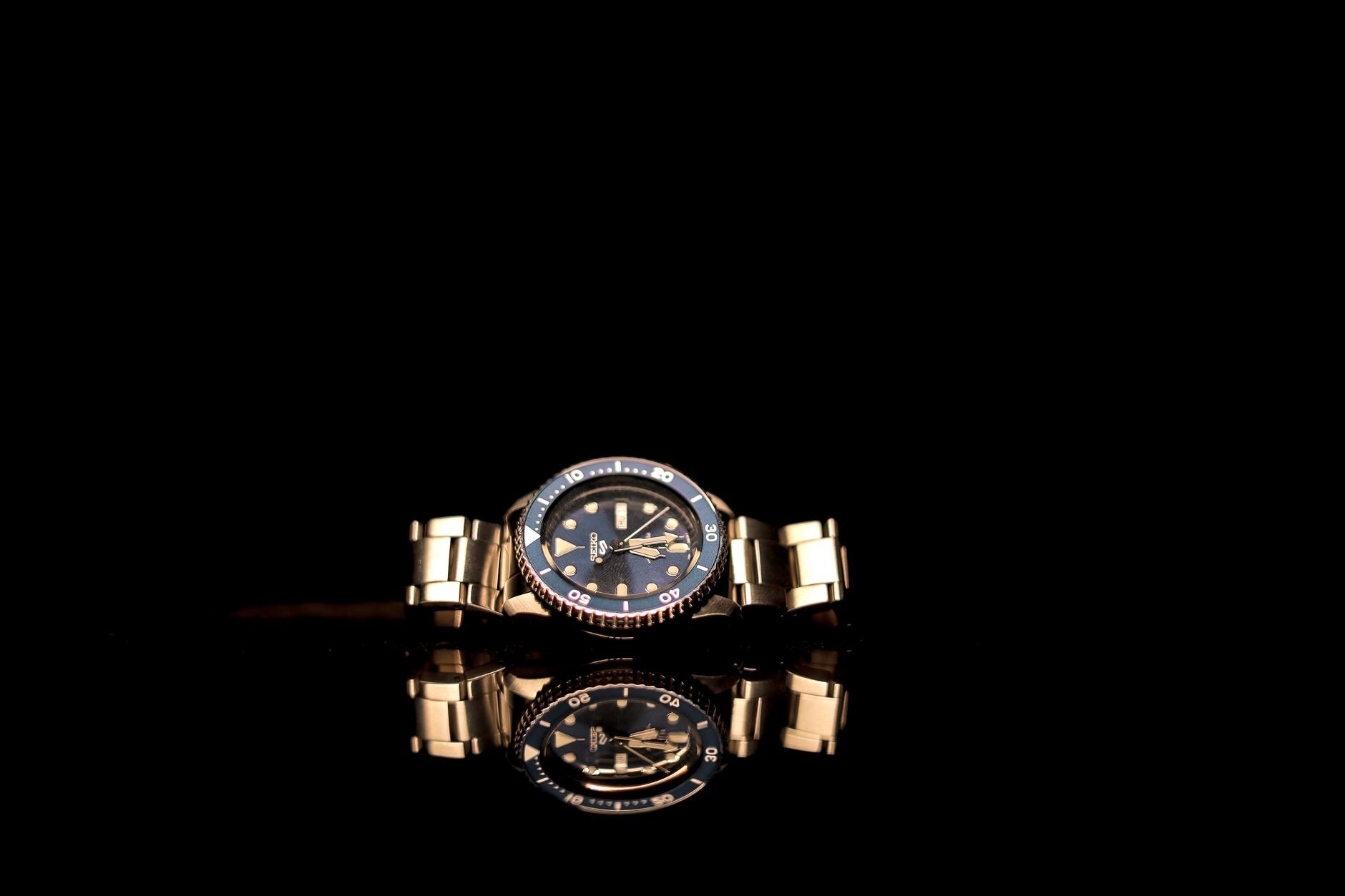 A luxurious gold wristwatch with a black dial beautifully reflected on a glossy surface.