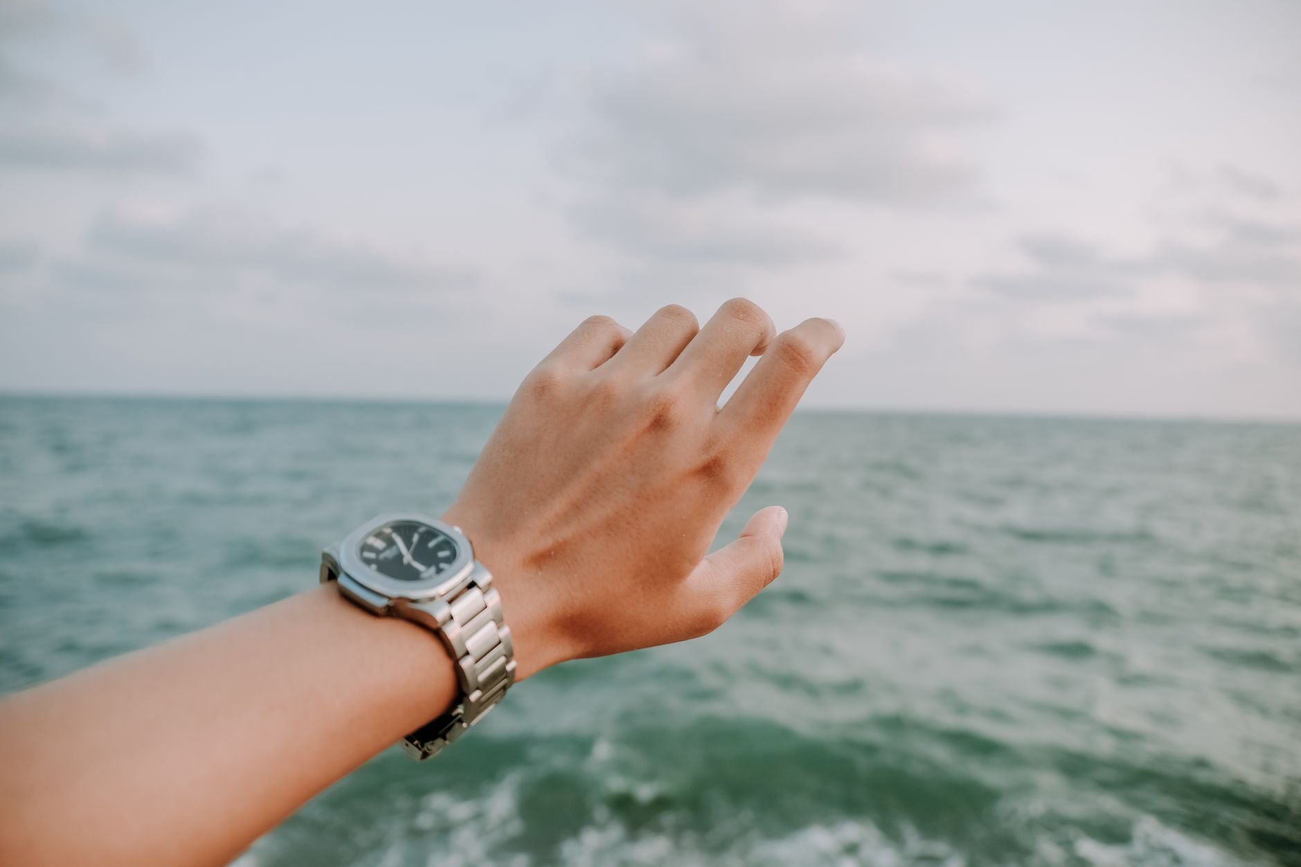 A close-up shot of a hand wearing a wristwatch, reaching towards the ocean under a cloudy sky.