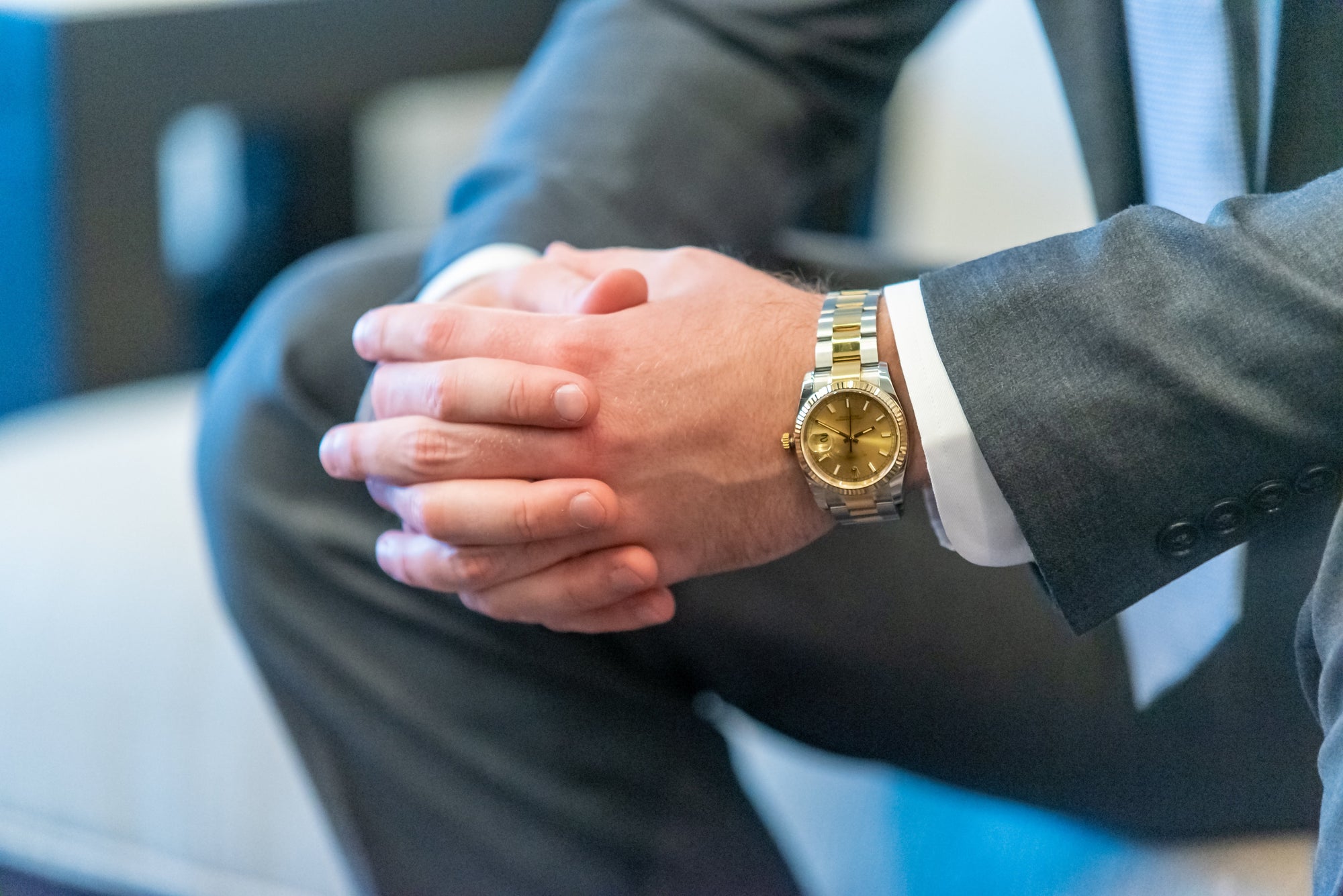 Close-up of a person in a gray suit sitting with hands clasped, wearing a two-tone gold and silver wristwatch.